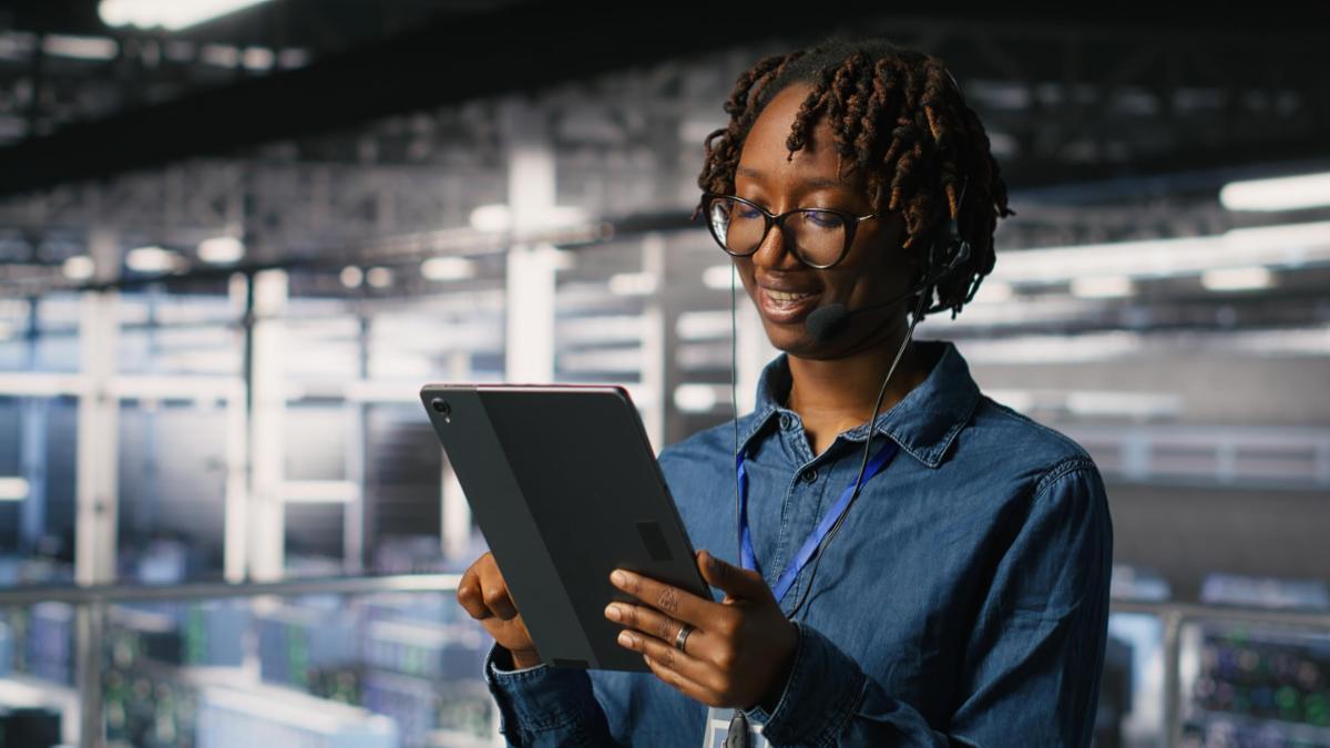 Data center engineer wearing headset microphone providing remote diagnostics using tablet. Technician monitors system health and guides users through infrastructure troubleshooting steps