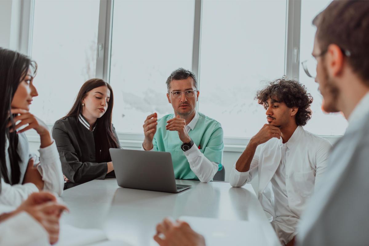 A group of multiethnic medical professionals including doctors, surgeons, and nurses are gathered in a hospital setting discussing patient care and using modern technology to address challenges in